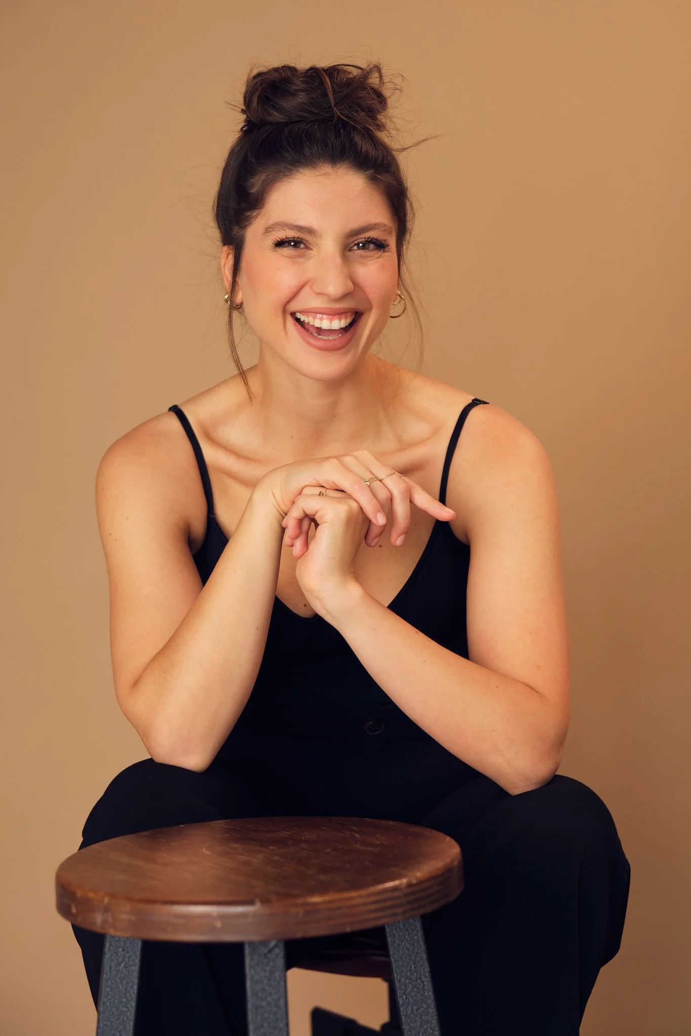 A woman with dark hair in a messy bun, wearing a black spaghetti strap top, smiling and looking at the camera, sitting behind a wooden stool against a beige background.