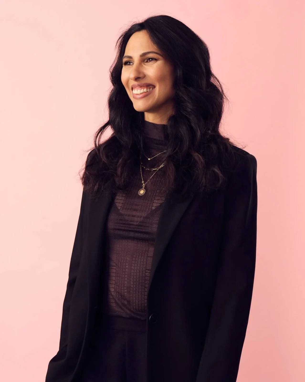 A woman with long black hair smiling, wearing a black blazer and jewelry, standing against a pink background.