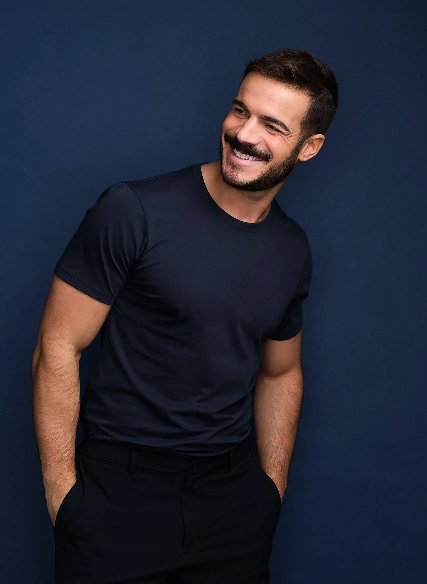 A smiling man with dark hair and beard, wearing a dark blue t-shirt, standing against a dark blue background.