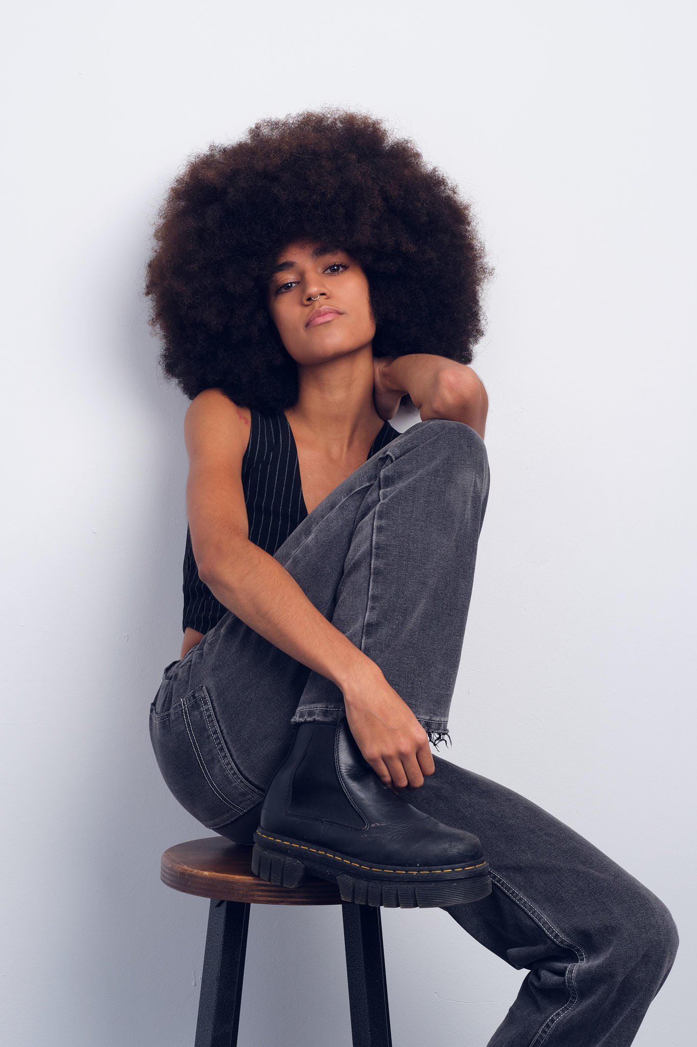 A woman with a large curly afro hairstyle, sitting on a stool against a plain white wall, wearing a black sleeveless top, gray jeans, and black boots.