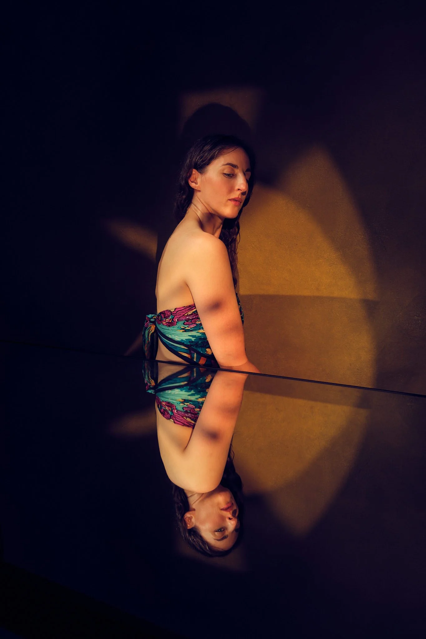 A woman with long brown hair wearing a multicoloured top posing pensively above a mirror against a mottled textured background