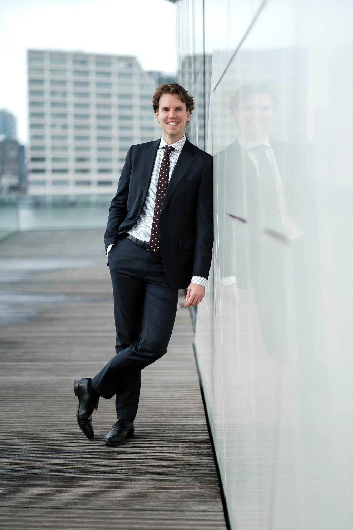 A businessman in a suit leaning against a glass wall on an outdoor balcony or rooftop, with city buildings in the background.