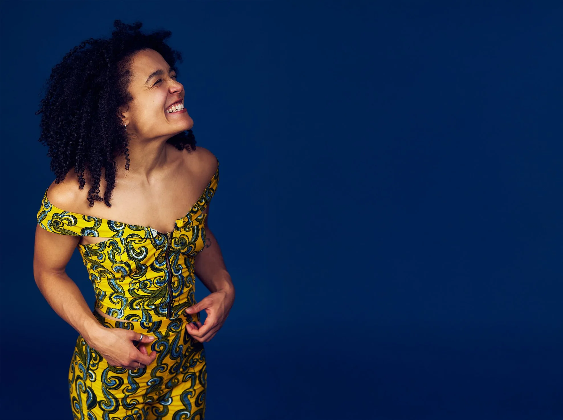 A woman with black curly shoulder length hair wearing a yellow with swirls two piece off the shoulder outfit standing and smiling with eyes closed against a dark blue background