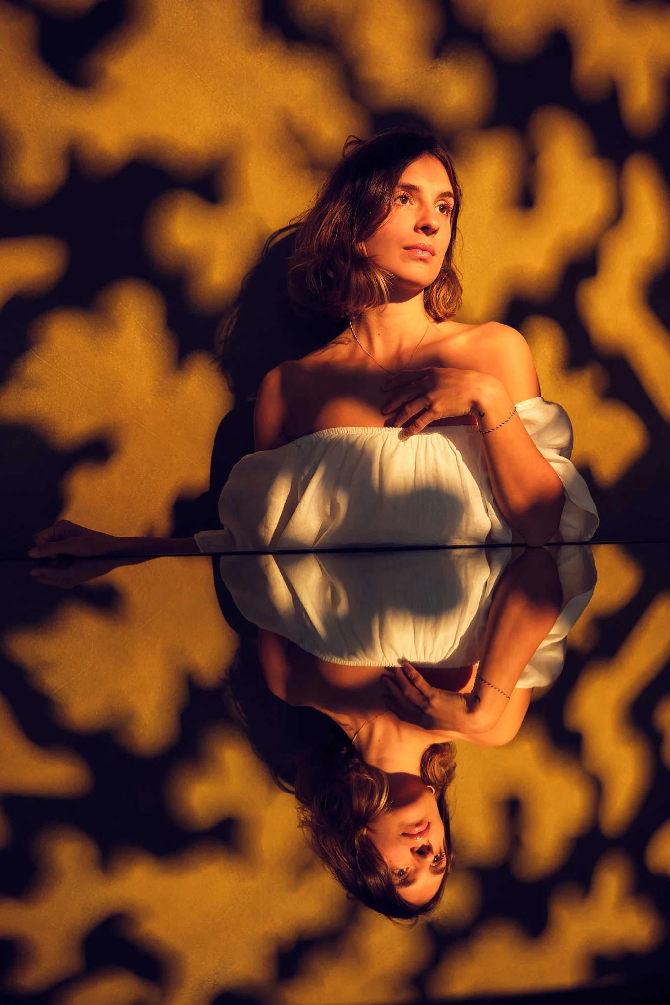 A woman wearing an off-the-shoulder white top with one hand resting on her chest posing above a mirror against a dappled dark yellow background