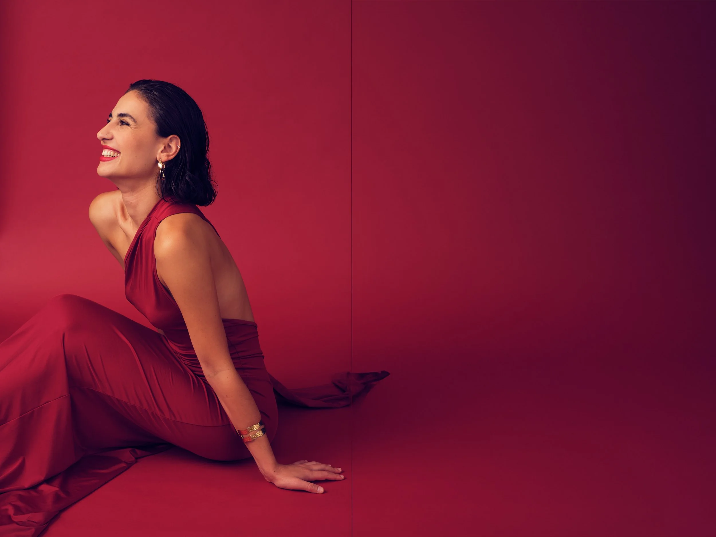Smiling woman in a red satin dress sitting on the floor against a red background.