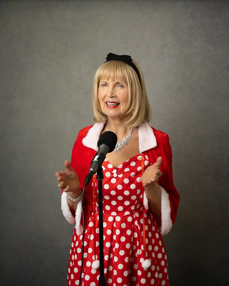 Jackie Mitchell as Doris Daydream in a red polka dot dress with a white collar and a red jacket with white trim, standing in front of a microphone. She has blonde hair, a black headband, and is wearing a sparkling necklace and bracelet. She appears t