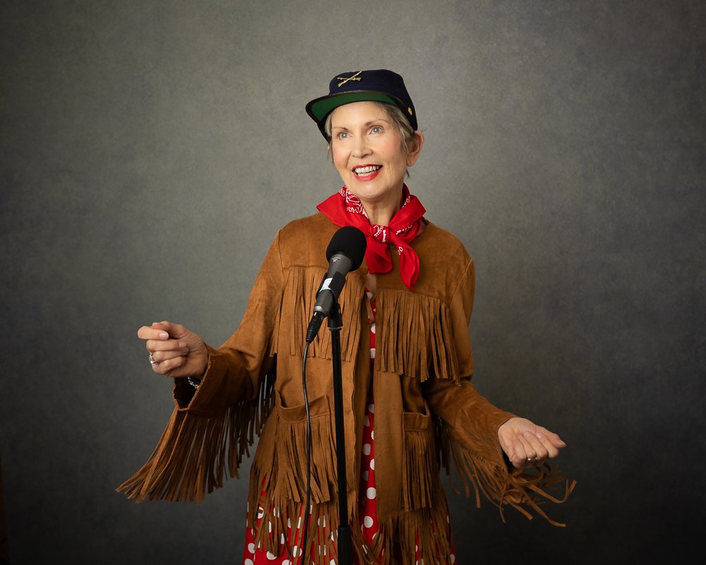 Jackie Mitchell wearing a brown fringed leather jacket, a red polka dot scarf, a black baseball cap, and a red bandana around her neck, standing in front of a microphone against a gray background.