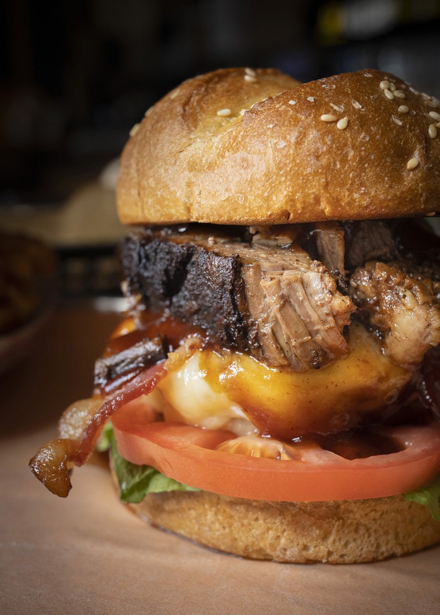 Close-up of a cheeseburger with a sesame seed bun, lettuce, tomato, bacon, beef brisket, and a beef patty.
