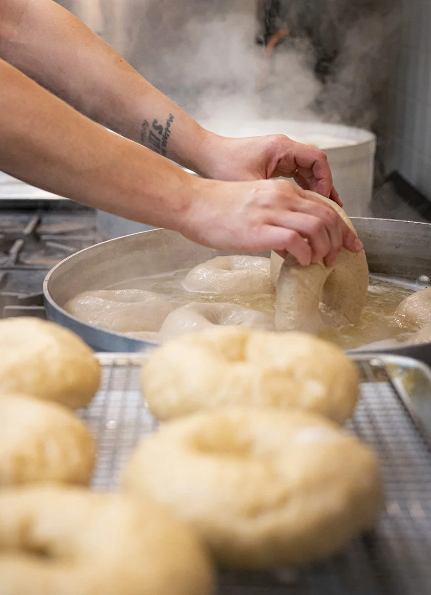 A person making bagels in boiling water, with fresh bagels on a cooling rack in the foreground.