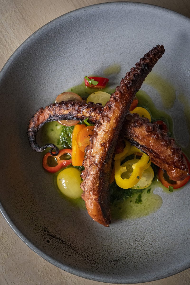 Grilled octopus tentacles served over a colorful salad with cherry tomatoes and yellow bell peppers in a dark bowl.
