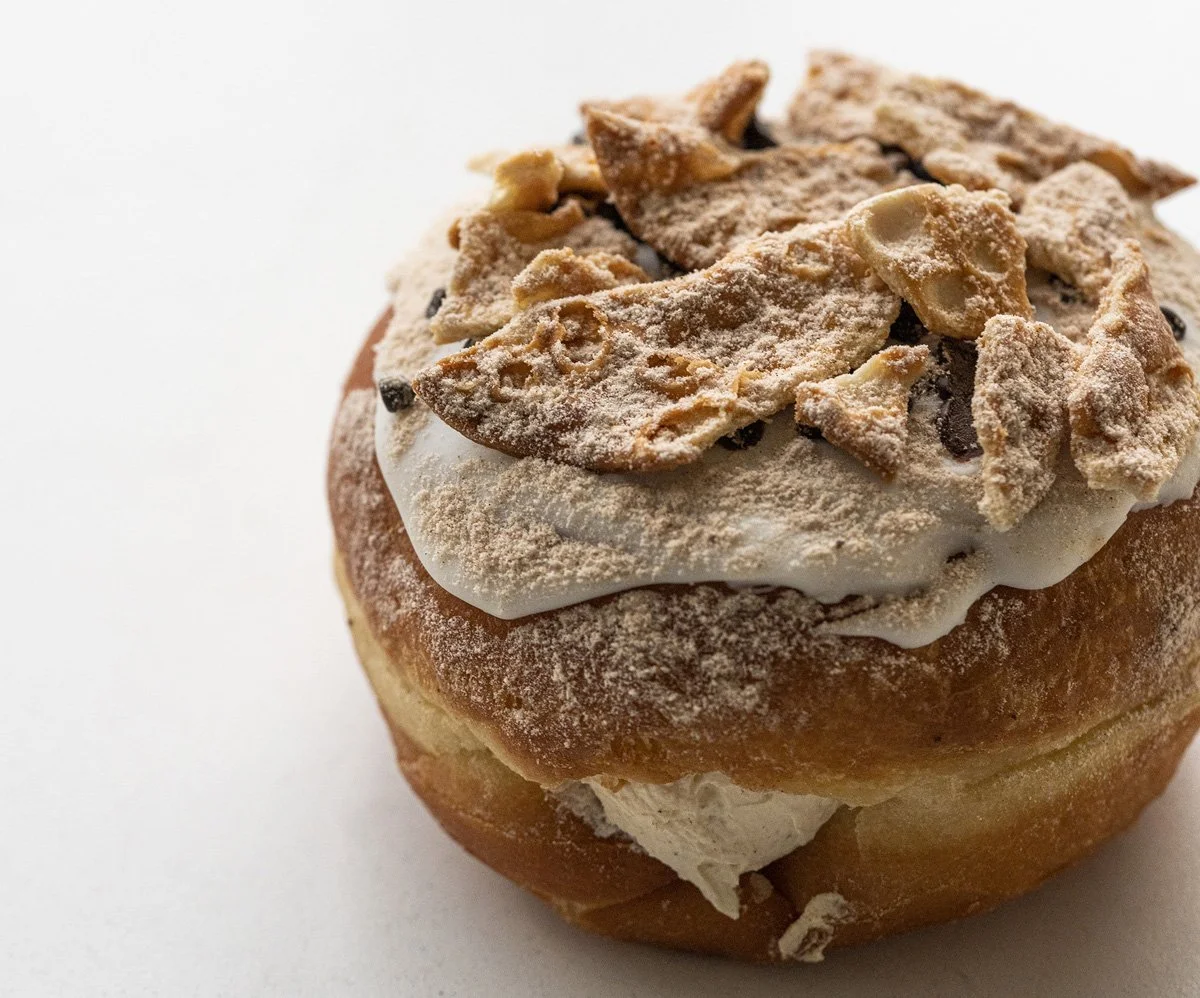 Close-up of a cannoli doughnut with creamy filling topped with crushed cannoli shell pieces and powdered sugar on a white background.