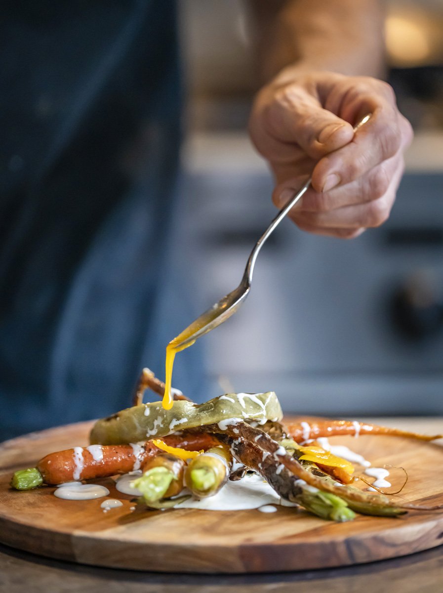 A chef's hand drizzling yellow sauce over roasted vegetables on a wooden plate.