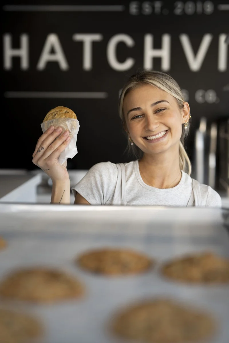 A smiling woman holding a cookie in her right hand inside a bakery named Hatchville.