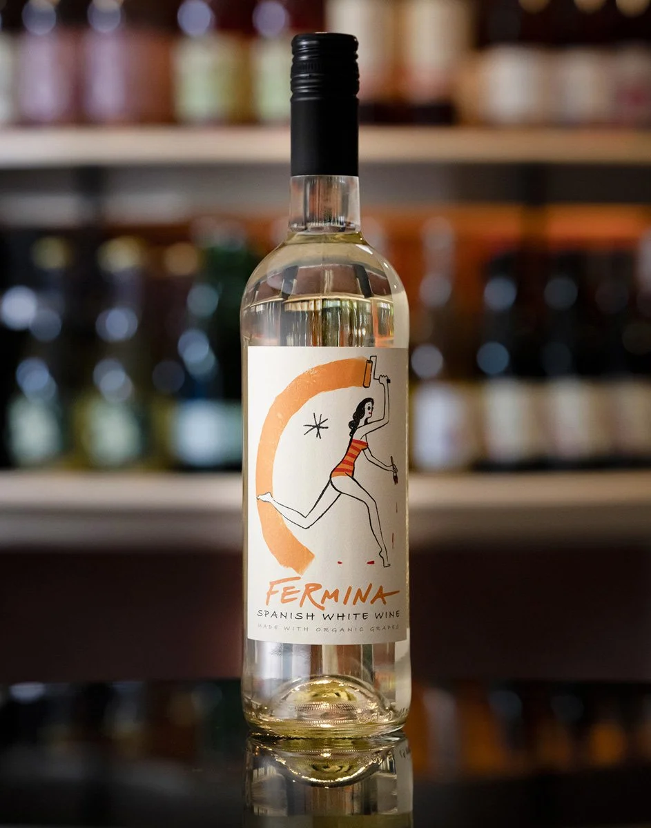 A bottle of Ferina Spanish white wine with a label featuring a stylized illustration of a woman in a striped swimsuit holding a hammer and standing on one leg, against a blurred background of wine bottles on shelves.