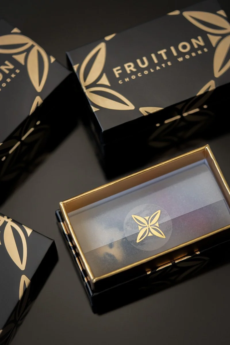 Black and gold packaging box with the label "Fruition Chocolate Works" and a floral logo, partially open revealing paper with a sticker inside.