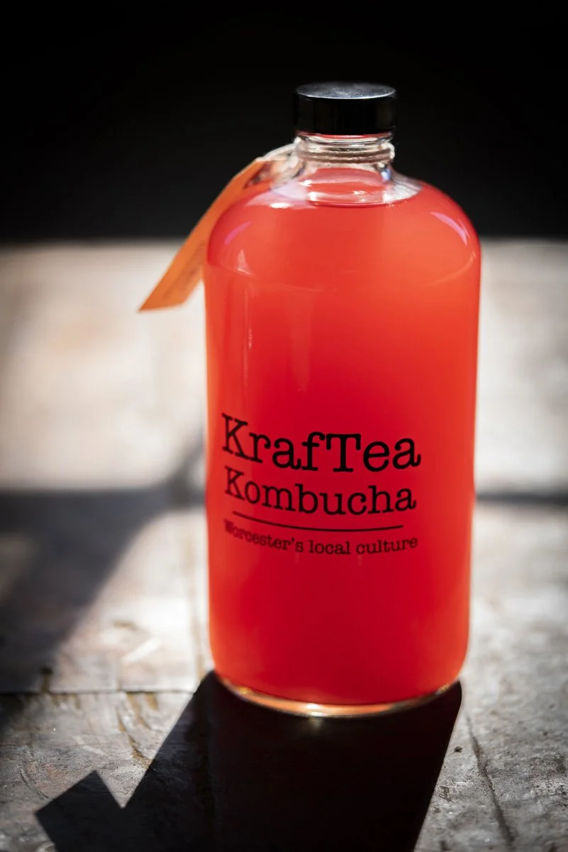 A glass bottle with a black cap containing reddish-pink liquid labeled "KrafTea Kombucha", placed on a wooden surface with a dark background.