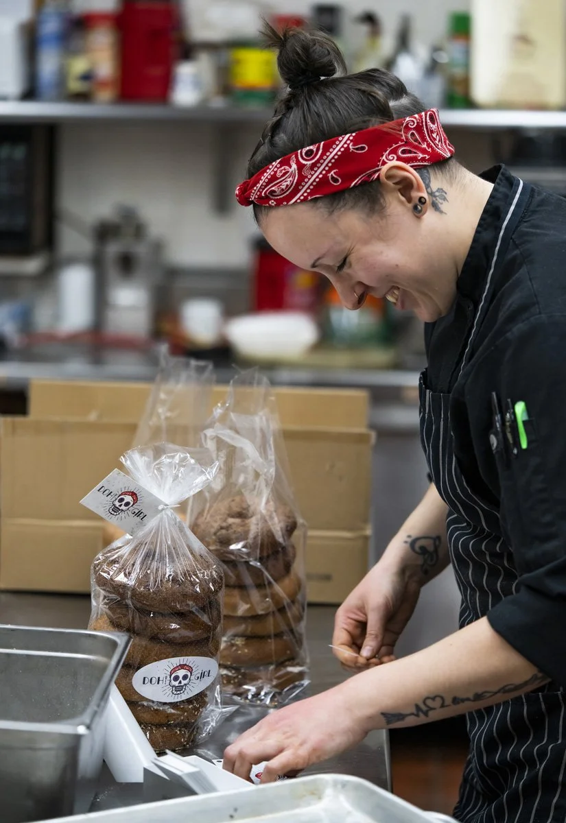 A woman with tattoos and a red bandana packaging bagels in a kitchen, with decorated bagel bags labeled "DohGirl" on the front.