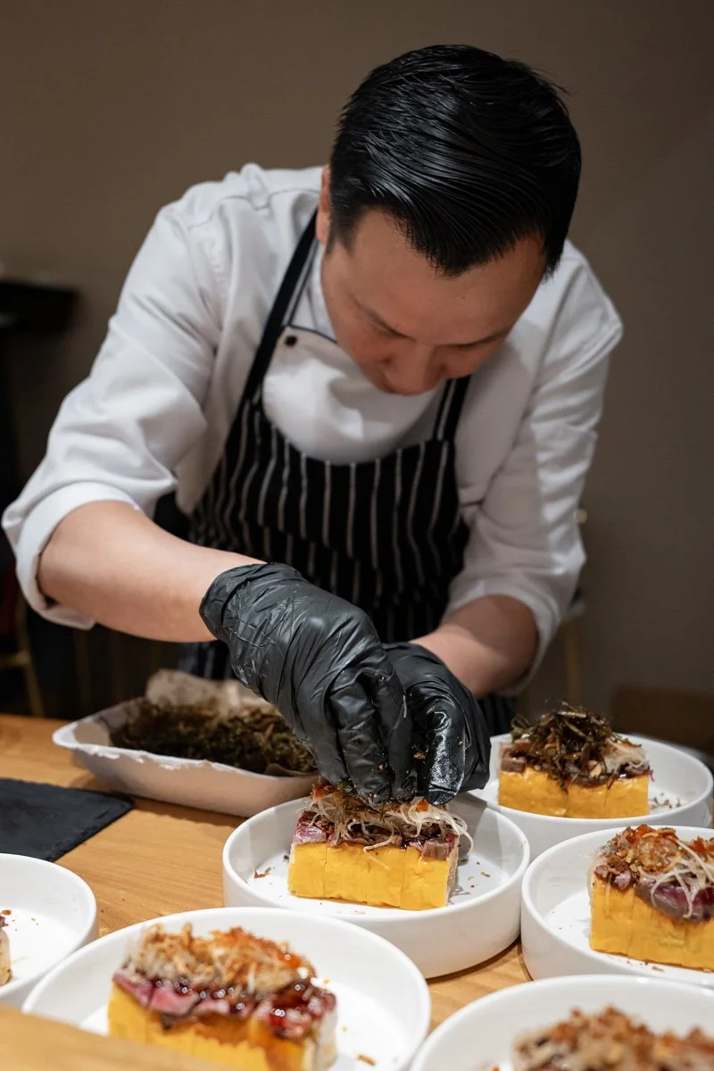 Chef in a white uniform and striped apron garnishes sushi rolls with seared sliced beef and toppings in white bowls.