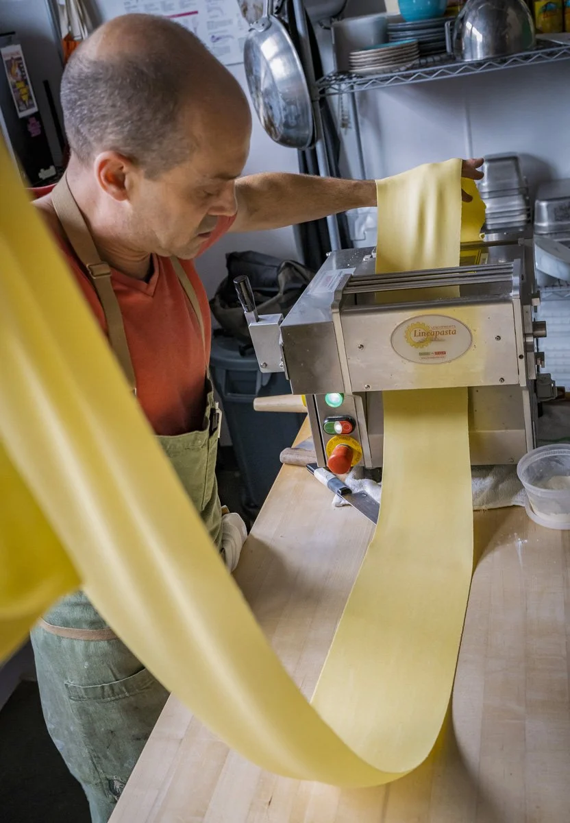 A man operating a pasta machine, rolling out fresh pasta dough in a kitchen.