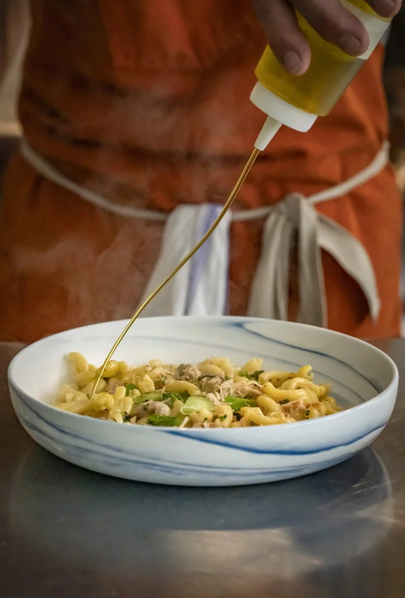 Person drizzling olive oil onto a bowl of pasta with vegetables.