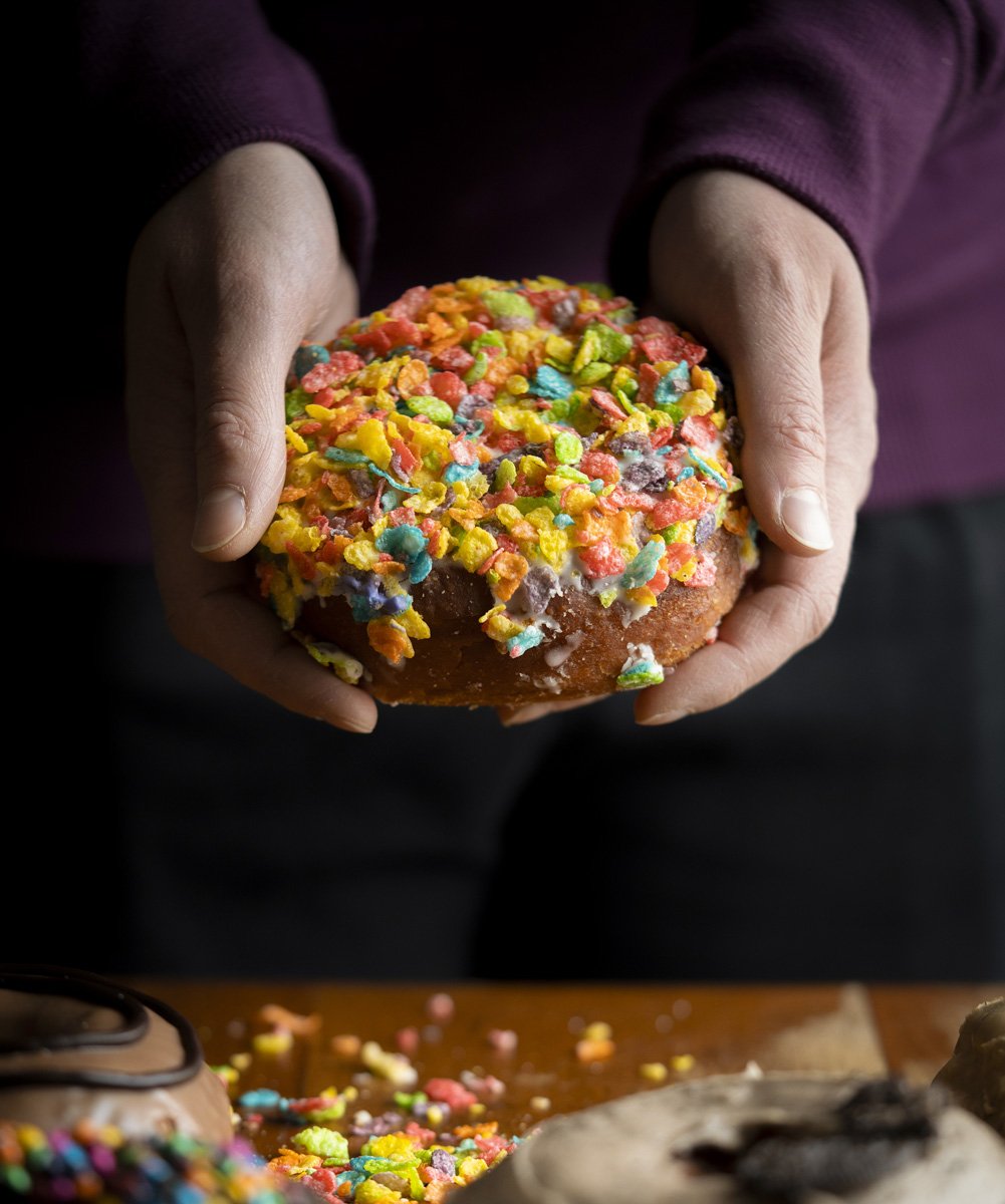 Person holding a doughnut topped with colorful Fruity Pebbles cereal.