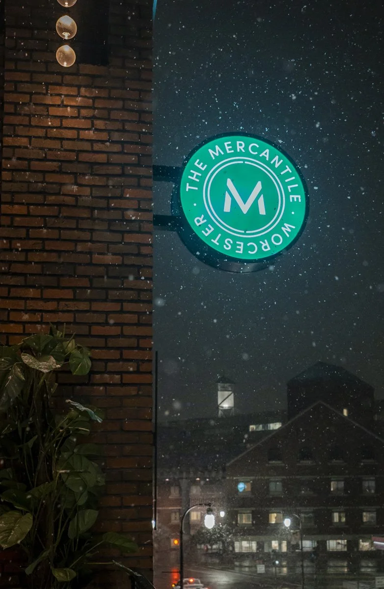 Nighttime street scene with a neon sign reading 'The Mercantile' and 'Worcester' on a building, visible snow falling, and a brick wall with hanging light fixtures.