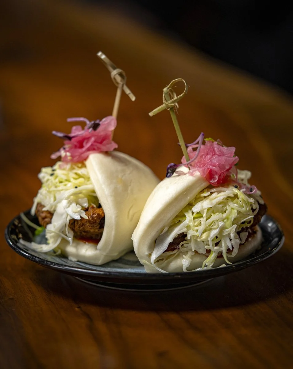 Two steamed pork buns filled with shredded cabbage, topped with pink pickled ginger and skewers, served on a black plate.