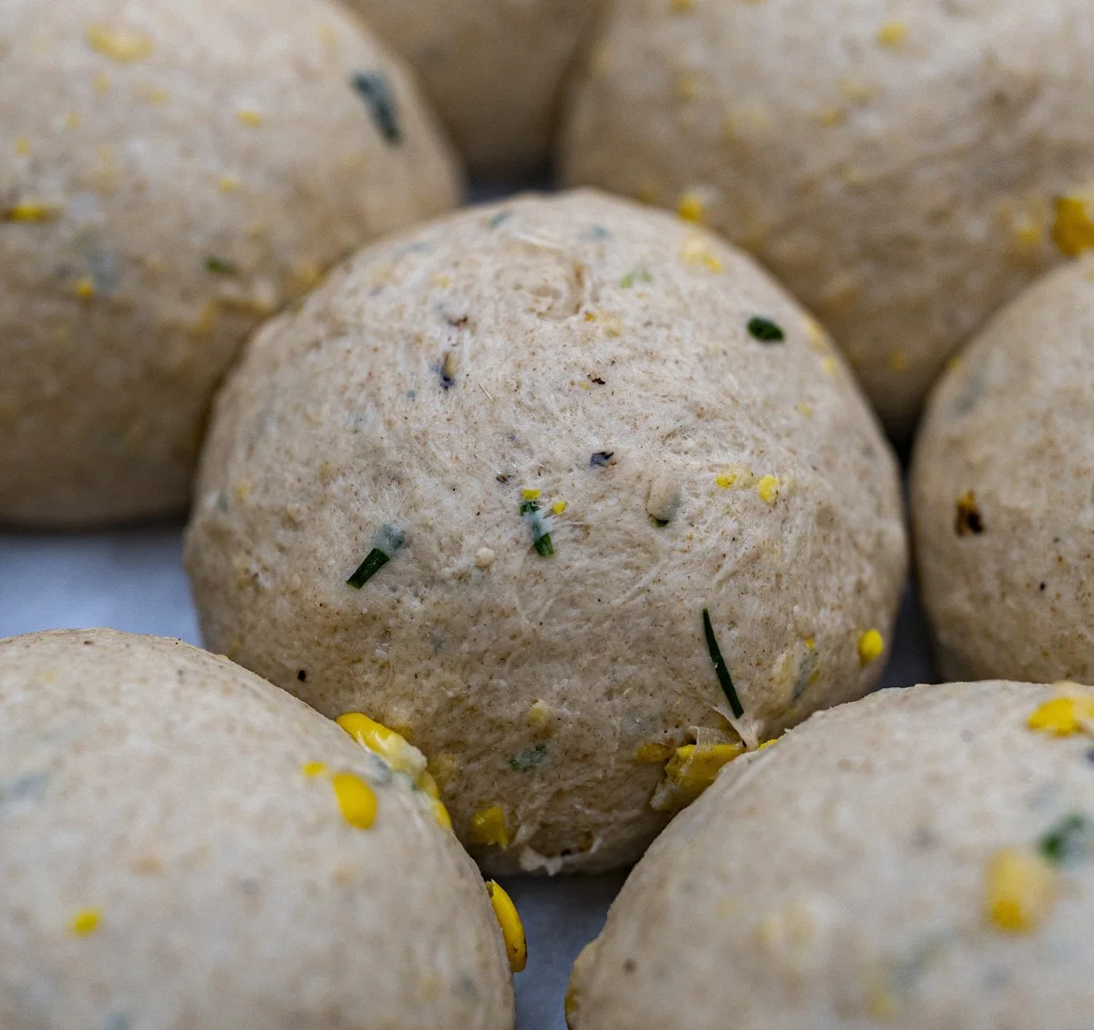 Close-up of a raw bagel dough ball with scallions and corn pieces, surrounded by similar balls.