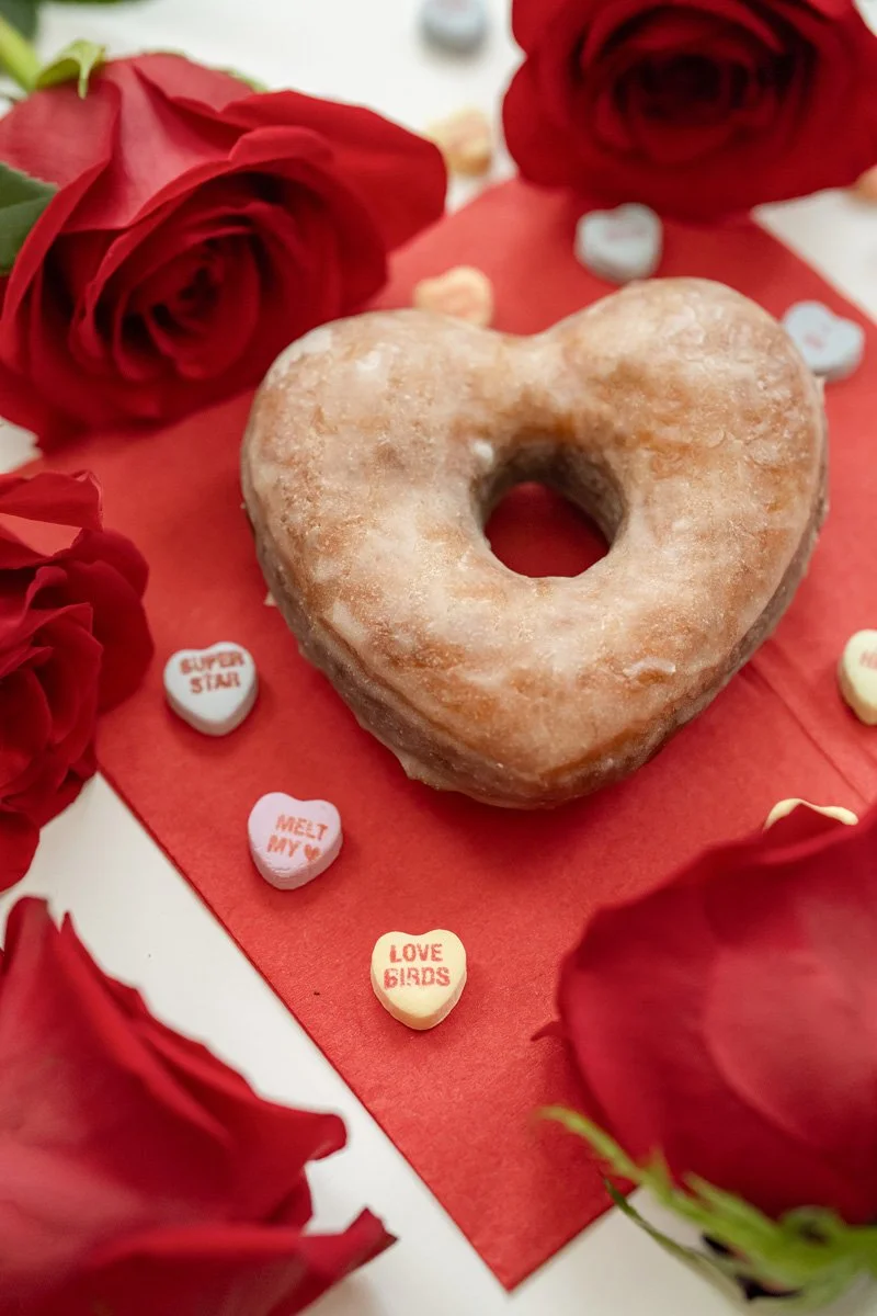 A heart-shaped donut surrounded by red roses and small conversation heart candies on a red and white background, celebrating love or Valentine's Day.