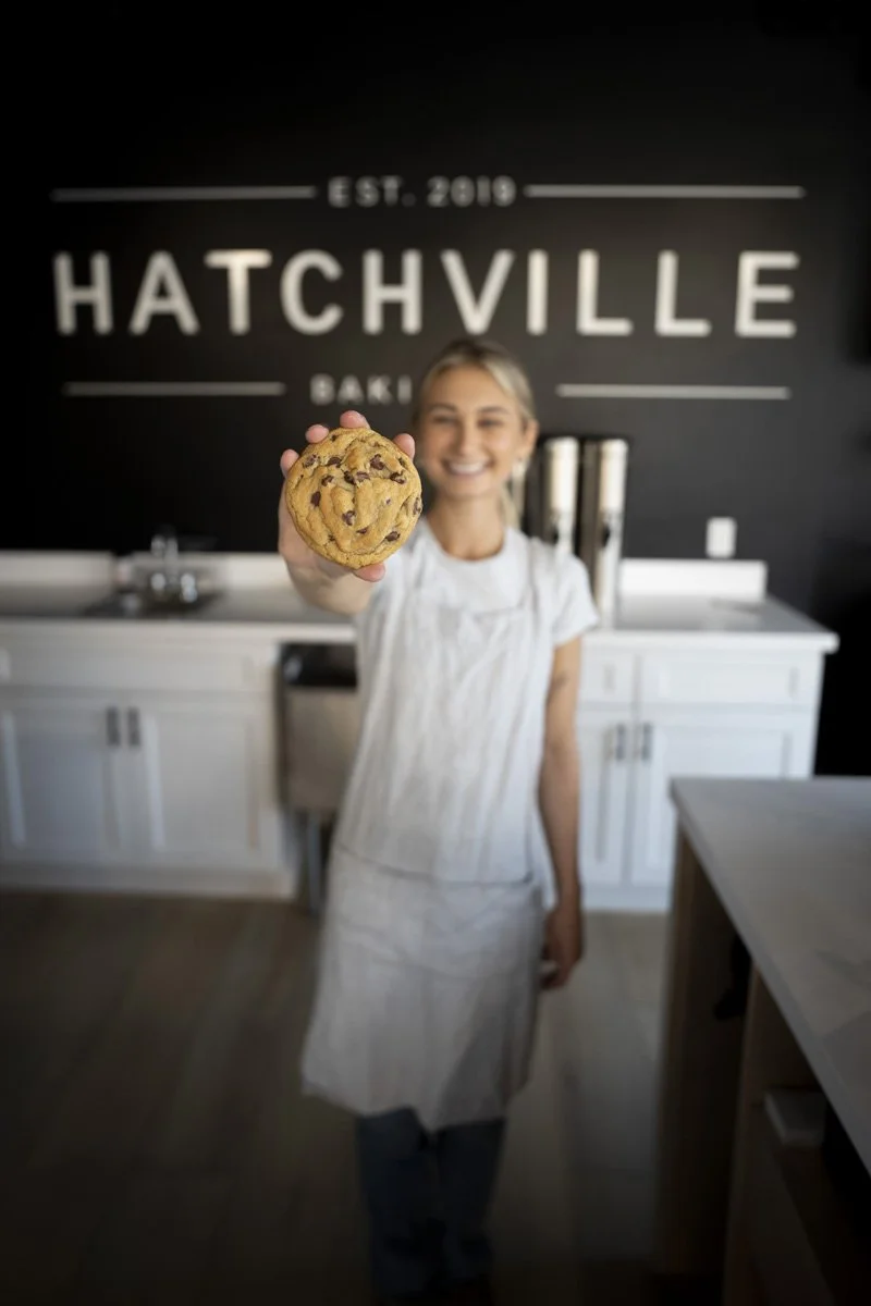Woman smiling and holding a chocolate chip cookie towards the camera in a bakery with a black wall and the sign 'Hatchville Bakery' in the background.