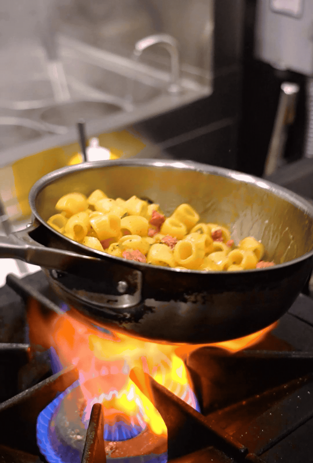 Cooking shells pasta with guanciale in a frying pan over an open flame on a stove.