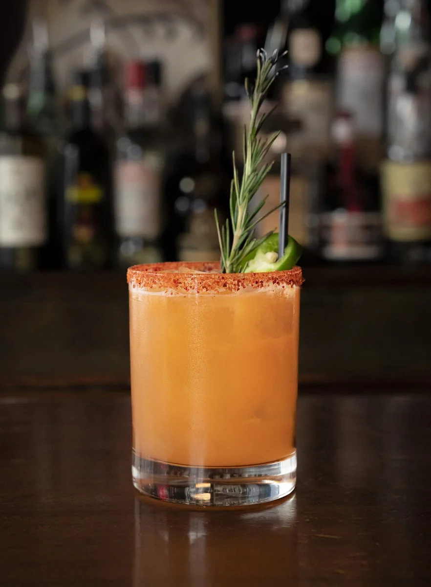 A cocktail in a clear glass with an orange-colored tequila drink and a tajin rim, garnished with a sprig of rosemary, a jalapeno slice, and a black straw, set on a dark wooden bar.