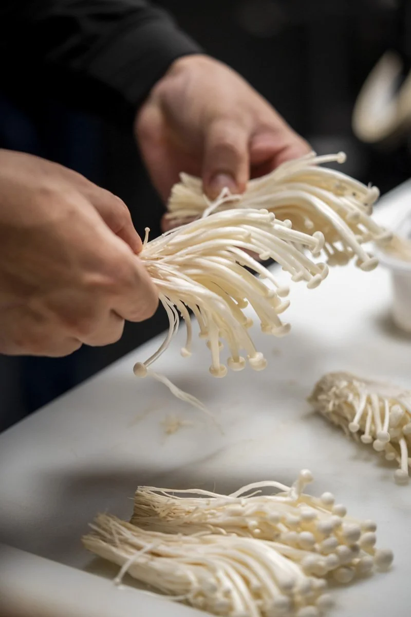 Person handling clusters of enoki mushrooms on a white surface.