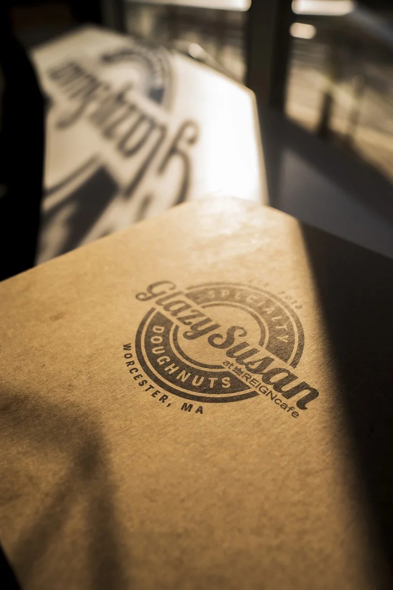 A doughnut box from Glazy Susan's in Worcester, MA, with a logo printed on it, resting on a surface with a blurred background.