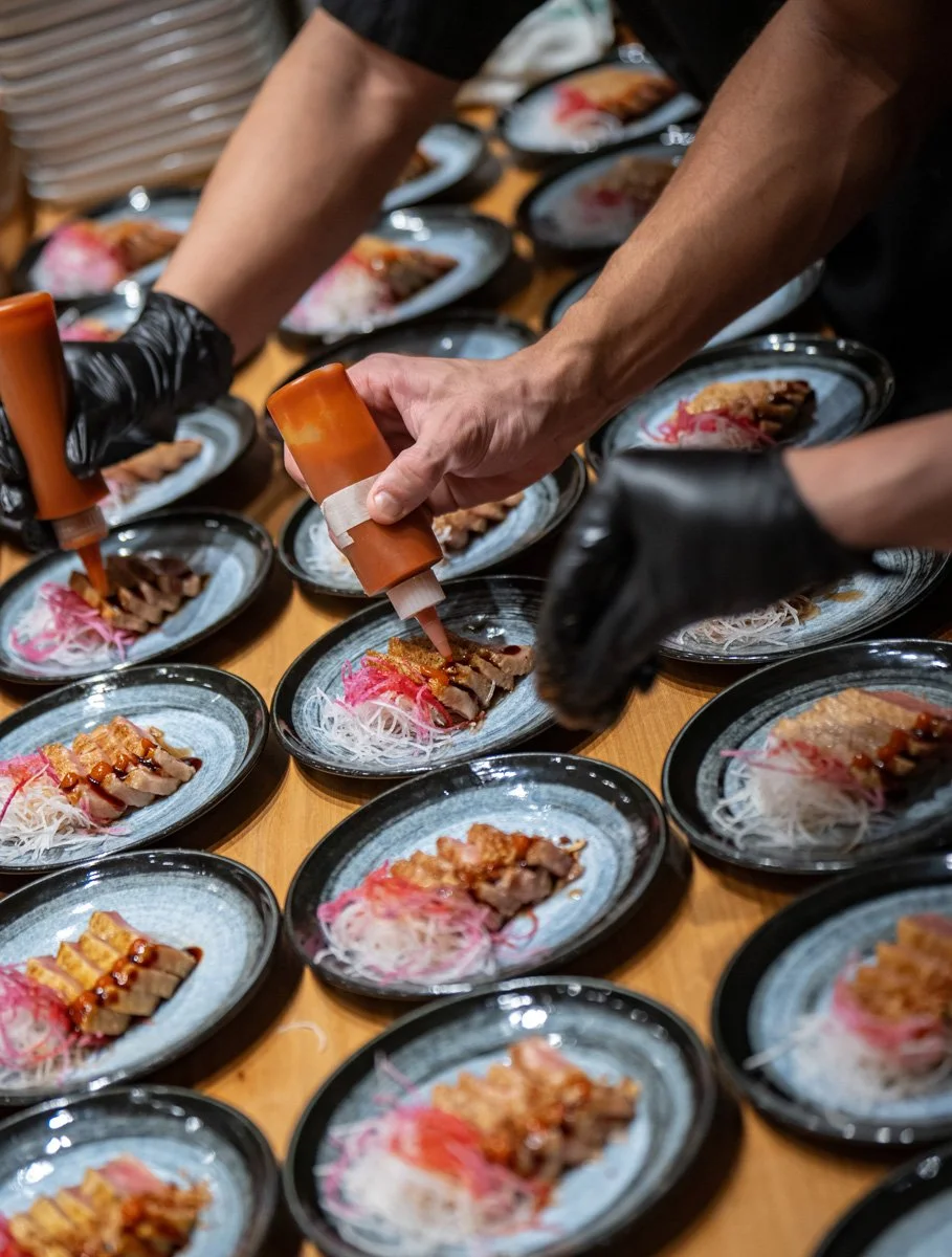 People preparing plates with sashimi, garnished with pink shredded radish, on a wooden table, using orange squeeze bottles for sauce.