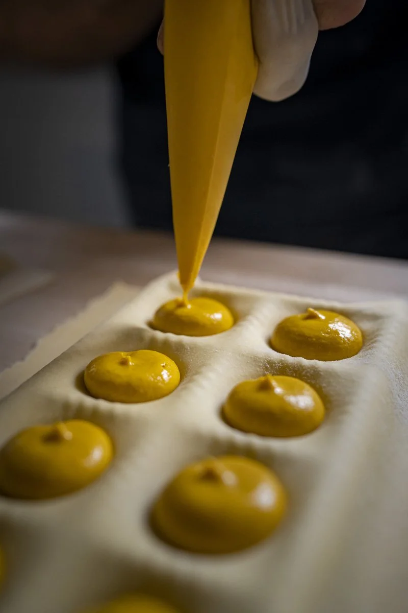 A chef's hand piping yellow filling into raw pasta wells to make ravioli.