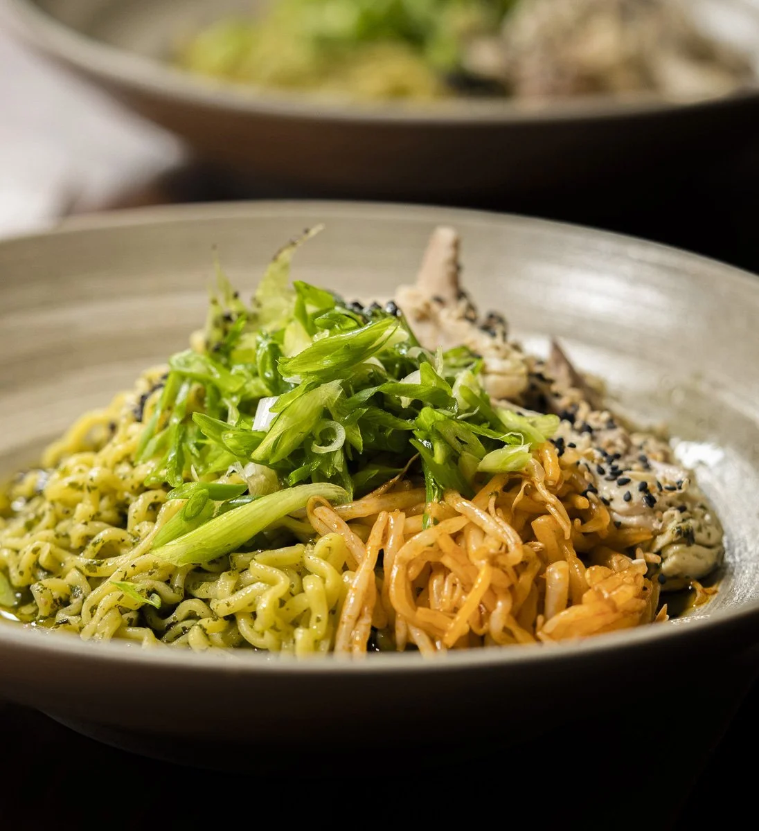 A bowl of Asian-inspired noodles topped with chopped green onions, sesame seeds, and slices of cooked chicken.