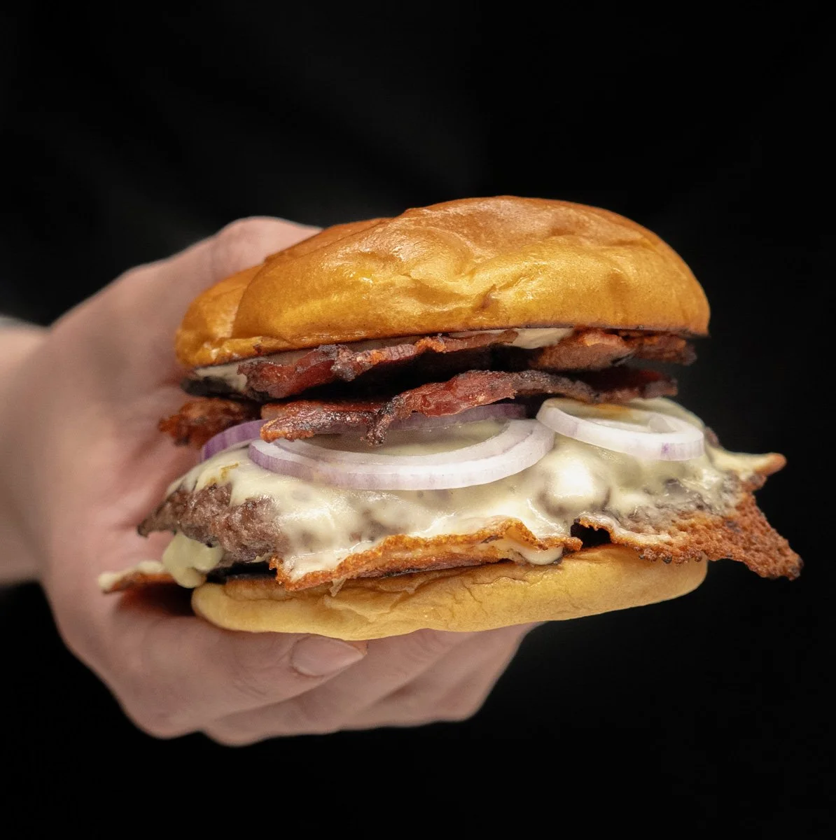 A close-up of a hand holding a bacon cheeseburger with onion slices, melted cheese, and a toasted bun against a black background.