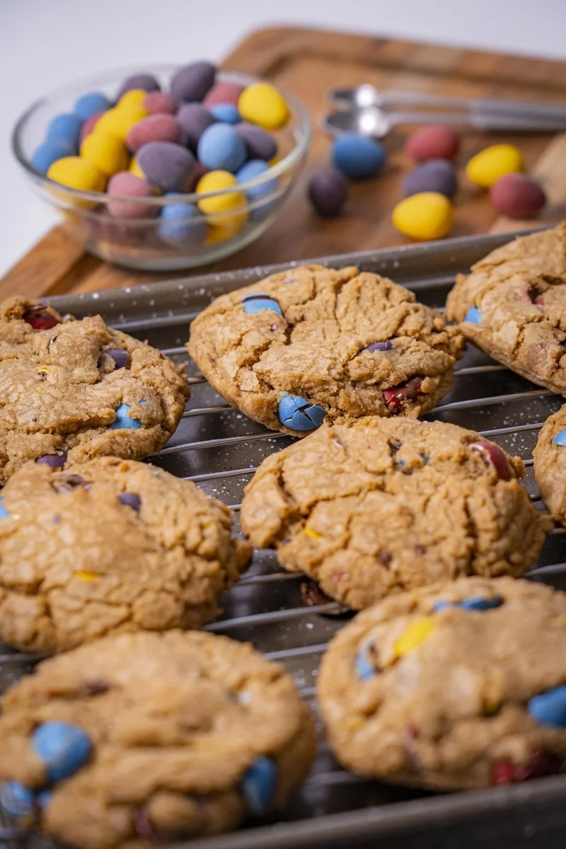 Freshly baked Easter cookies with colorful chocolate eggs cooling on a wire rack, with a bowl of extra chocolate eggs and a wooden cutting board in the background.