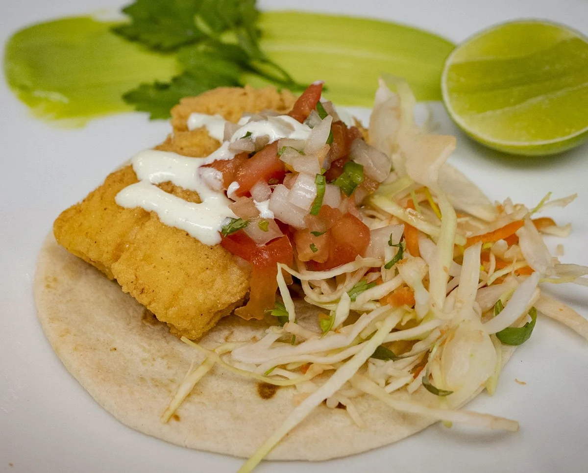 A fish taco on a soft tortilla, topped with shredded cabbage, pico de gallo, sour cream, and a drizzle of white sauce. Two lime wedges and a cilantro garnish in the background.