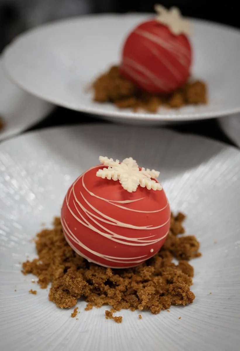A red spherical Christmas dessert decorated with white chocolate snowflake and lines, placed on a bed of crumbled cookie crumbs on a white plate.