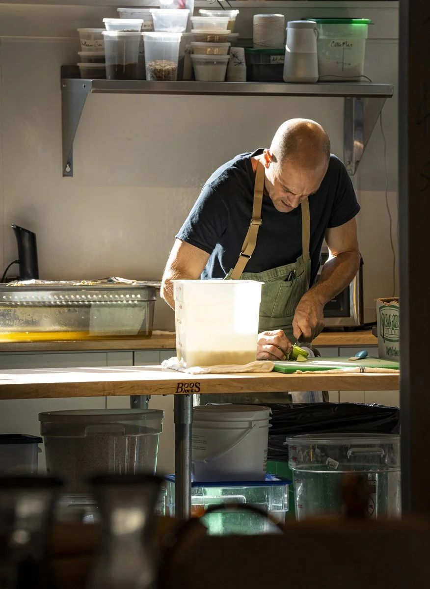 Man in black t-shirt and apron chopping green vegetables in a kitchen with containers of ingredients on shelves above and various kitchen supplies below.