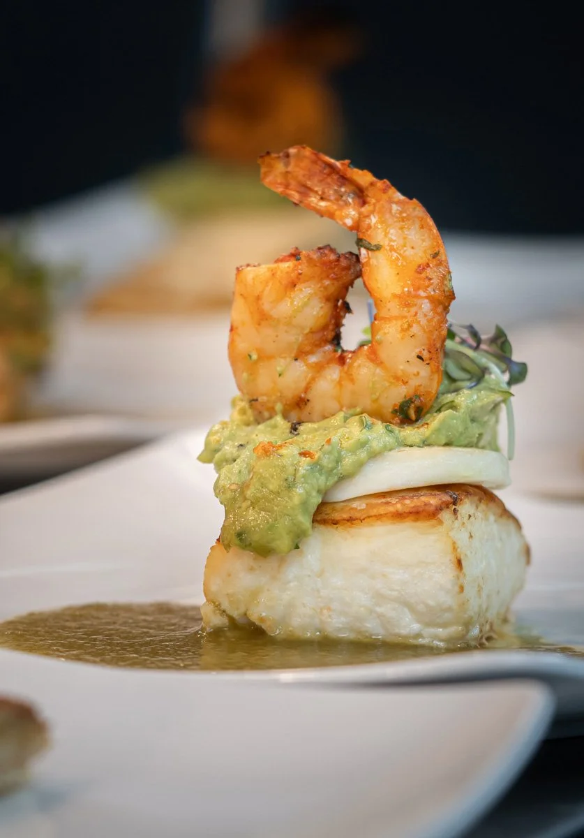 Close-up of a seafood dish with a grilled shrimp on top of fish, topped with guacamole and halloumi, with sauce underneath on a white plate.