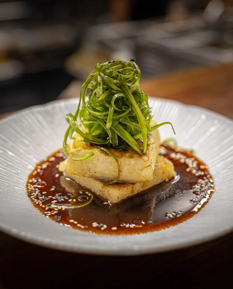 A plated gourmet dish featuring a piece of fish topped with seaweed garnish, served with a dark sauce and sesame seeds on a white textured plate.