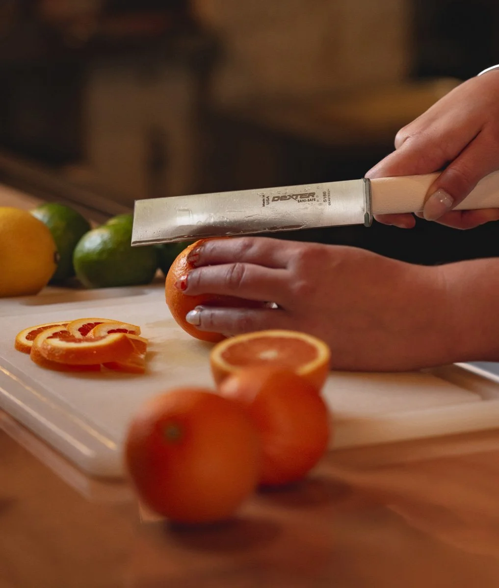 Person slicing an orange with a large kitchen knife, with whole citrus fruits and sliced oranges on a cutting board in a wooden bar setting.
