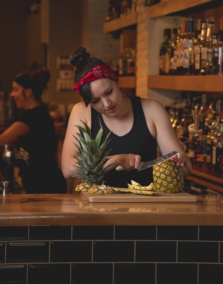 A woman with dark hair, a red bandana, and a black tank top slices a pineapple on a wooden bar counter. A pineapple with the top intact and pineapple peels are on the counter. In the background, another woman with her hair in a bun works behind the b