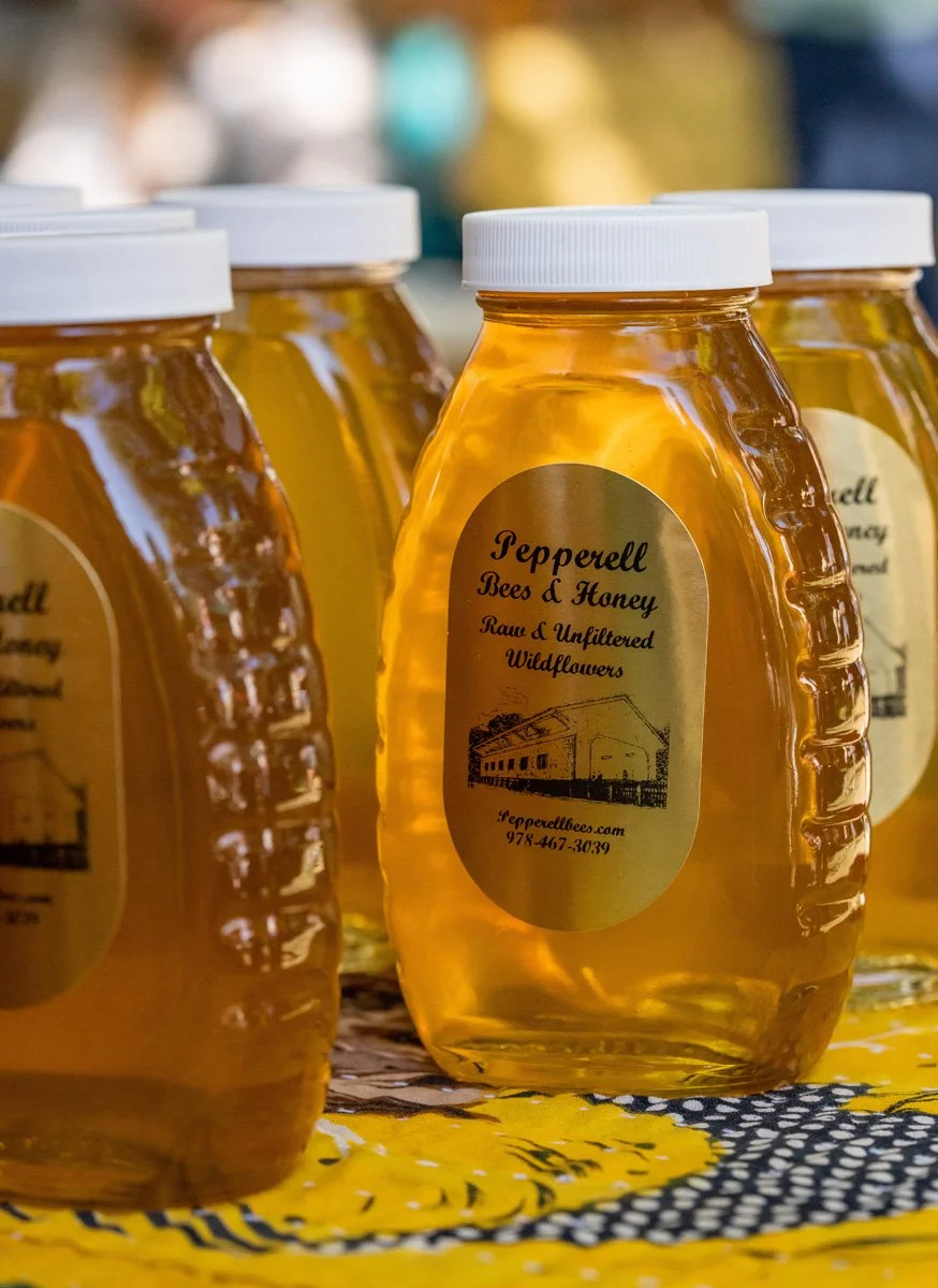 Jars of honey labeled 'Pepperell Bees & Honey, Raw & Unfiltered, Wildflowers' on a patterned yellow and black cloth.
