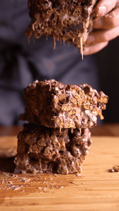 Stack of three cereal treats made out of Count Chocula on a wooden surface with a hand holding a piece above, dripping marshmallow and chocolate.