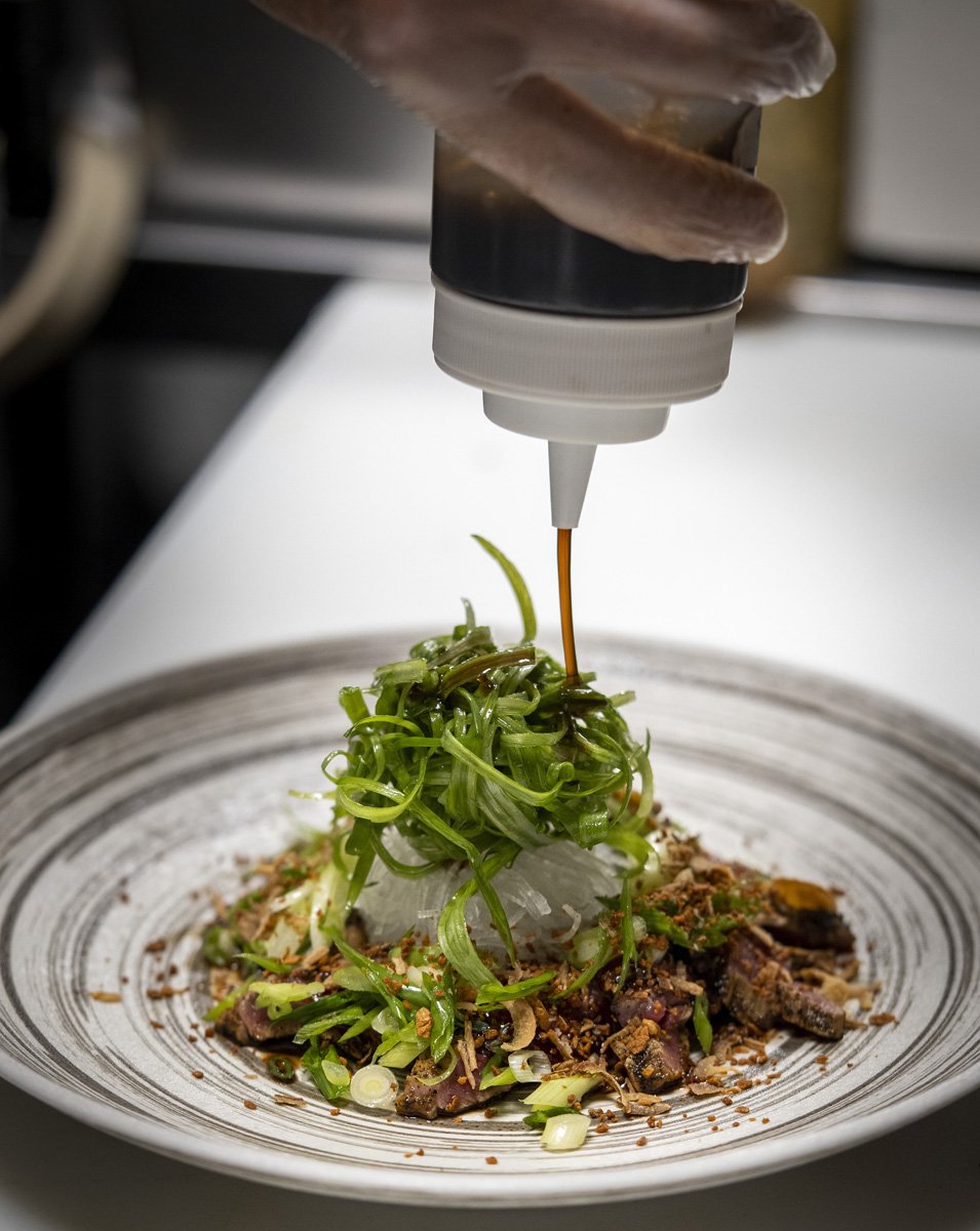 A chef drizzling sauce over a plated dish with sliced meat, green vegetables, and garnishes, on a white plate.