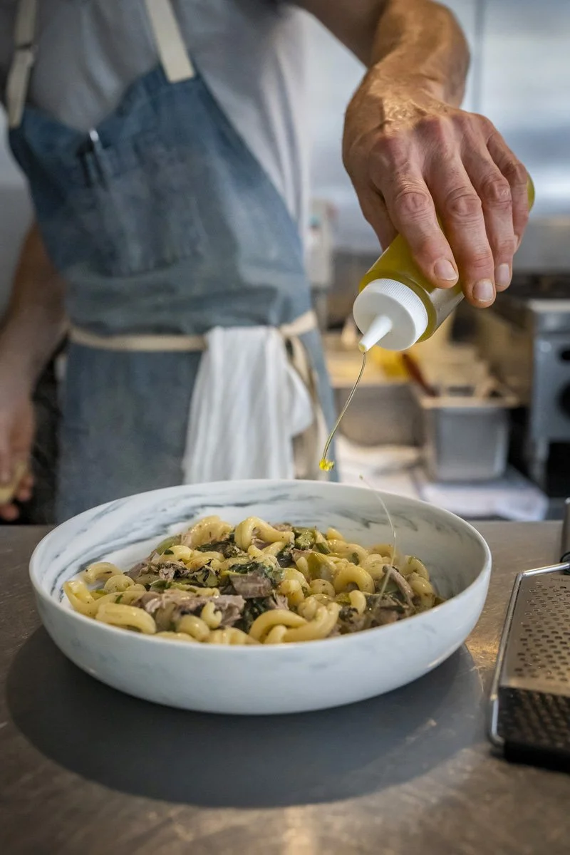 A person drizzling olive oil on a bowl of pasta with herbs and braised pork in a professional kitchen.